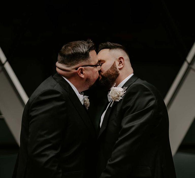 Grooms kissing in London wearing complimenting three piece black tuxedos with off white bow ties, white garden rose, lavender twig and dried foliage boutonniere and grooms Louboutins shoes 