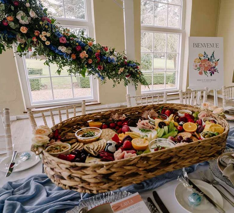 Large colourful wedding charcuterie board in wicker basket on classic wedding tablescape with white wedding tablecloth and cornflower blue wedding table runner