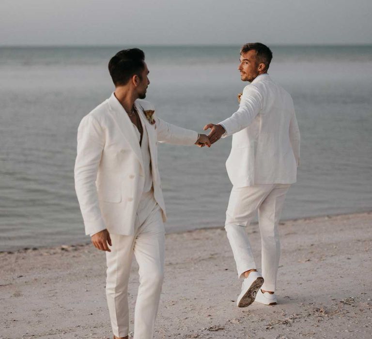 Grooms in complimenting double breasted white wedding suits holding hands doing couples photoshoot on the beach at Mexico destination wedding