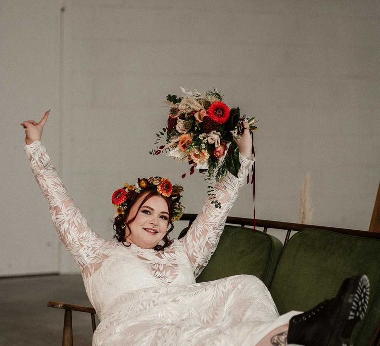 Bride in boho long sleeve 3D applique wedding dress with dried flower crown holding earthy toned garden rose, pampas grass, dried flower and foliage bridal bouquet sitting on crushed velvet olive green loveseat at The Barn at Avington