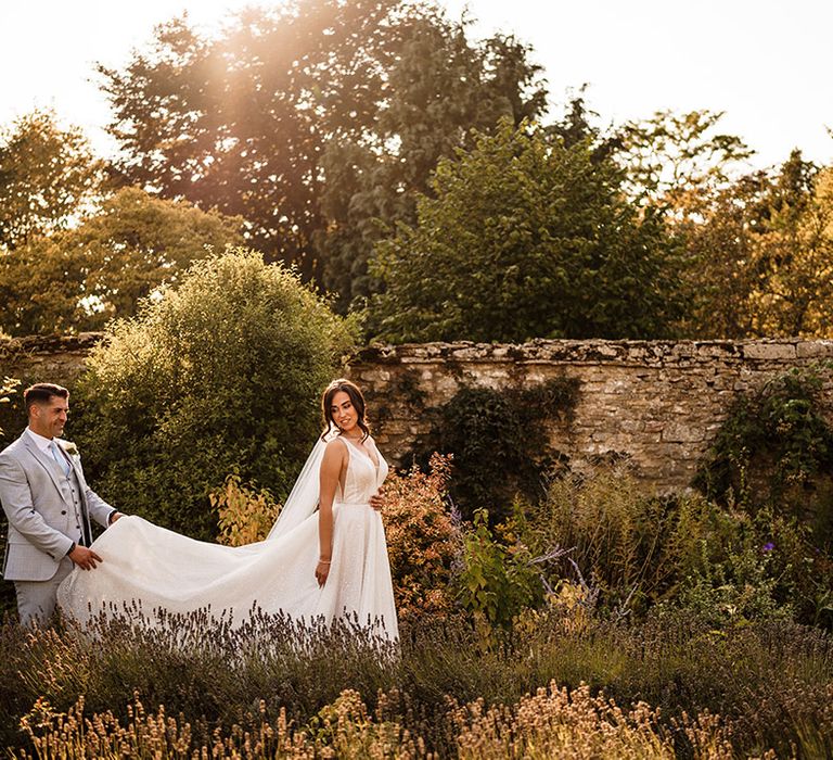 Groom in a light blue suit holding his brides wedding train in the gardens at Caswell House wedding venue in the Cotswolds 