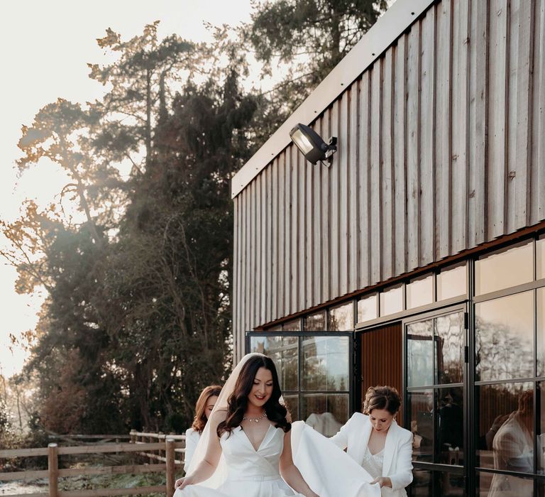 Bridesmaids in white bridesmaid suits follow bride in white silk wedding dress with thigh-high split