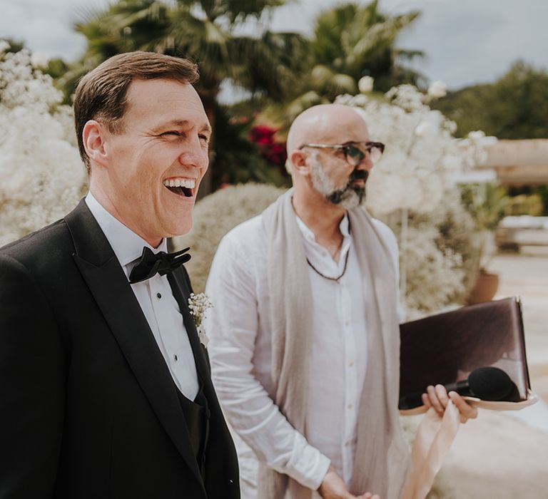 groom in a black tuxedo and bow tie smilling as his bride walks down the aisle at ibiza wedding