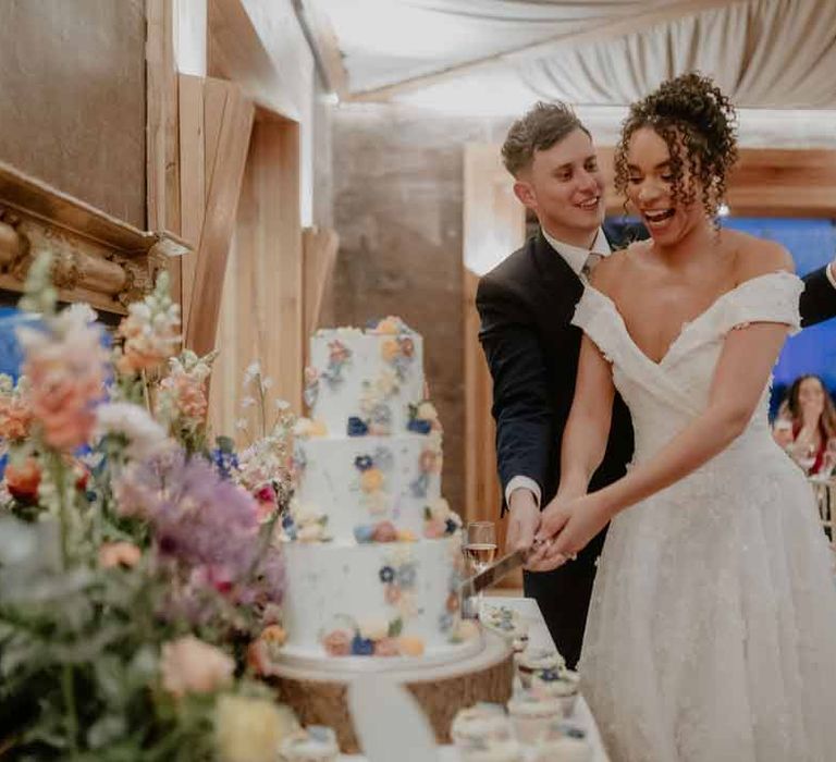 Groom in navy groom morning suit with grey ties and blush rose boutonniere standing with bride cutting the wedding cake in Suzanne Neville off shoulder 3D floral applique wedding dress