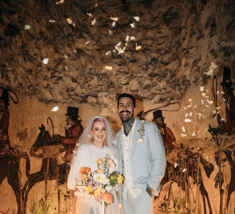 Bride in sheer overlay wedding dress and fluffy bridal coverup holding colourful bouquet with groom in baby blue linen suit, personalised embroidered linen shirt and garden rose and pampas grass boutonniere inside the Margate Caves