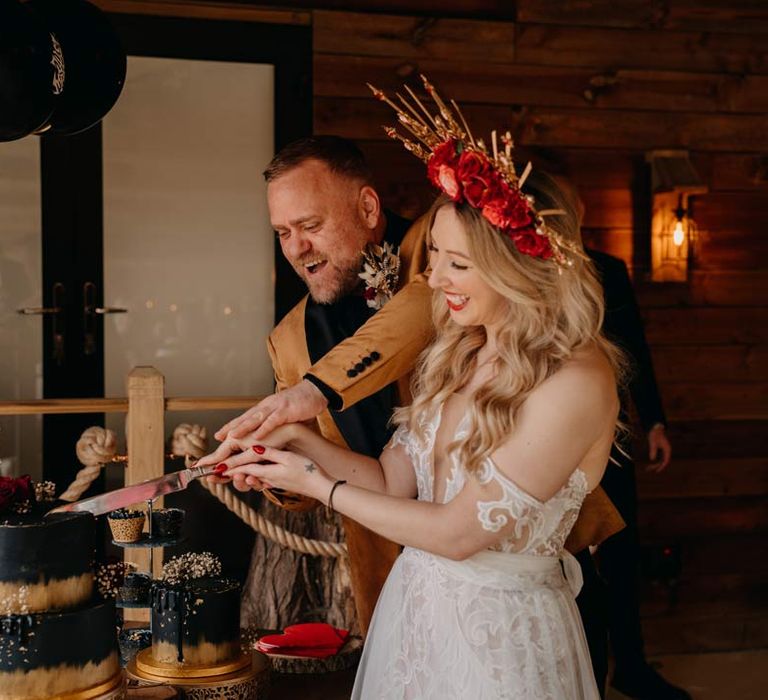 Groom in crushed velvet burnt orange grooms blazer and dried flower boutonniere cutting black wedding cakes with bride in off shoulder lace wedding dress with corset style top and red rose and gold halo bridal crown