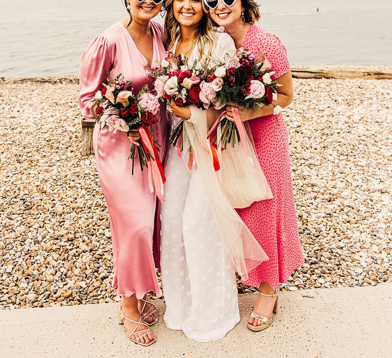 bridal party in heart sunglasses with wedding jumpsuit and different pink bridesmaid dresses 