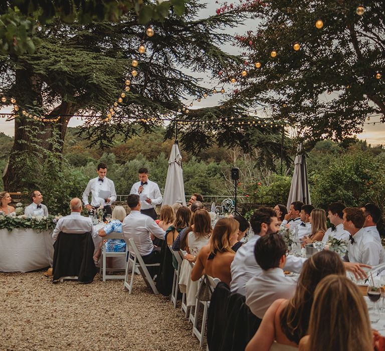 Outdoor destination wedding breakfast with festoon lighting and white minimalist decor 