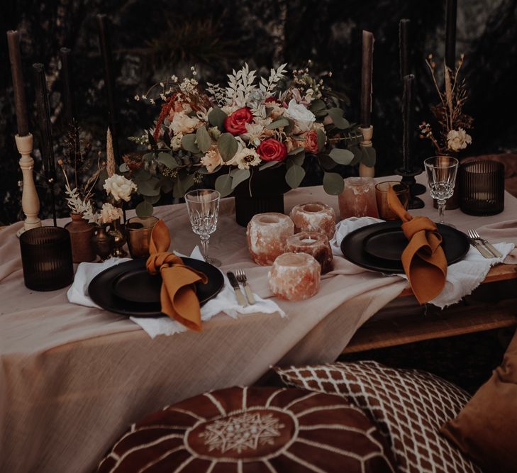 Table setting for two with black plates and orange ribbons next to Himalayan salt rock candle holders and black candles with a floral centrepiece of roses  