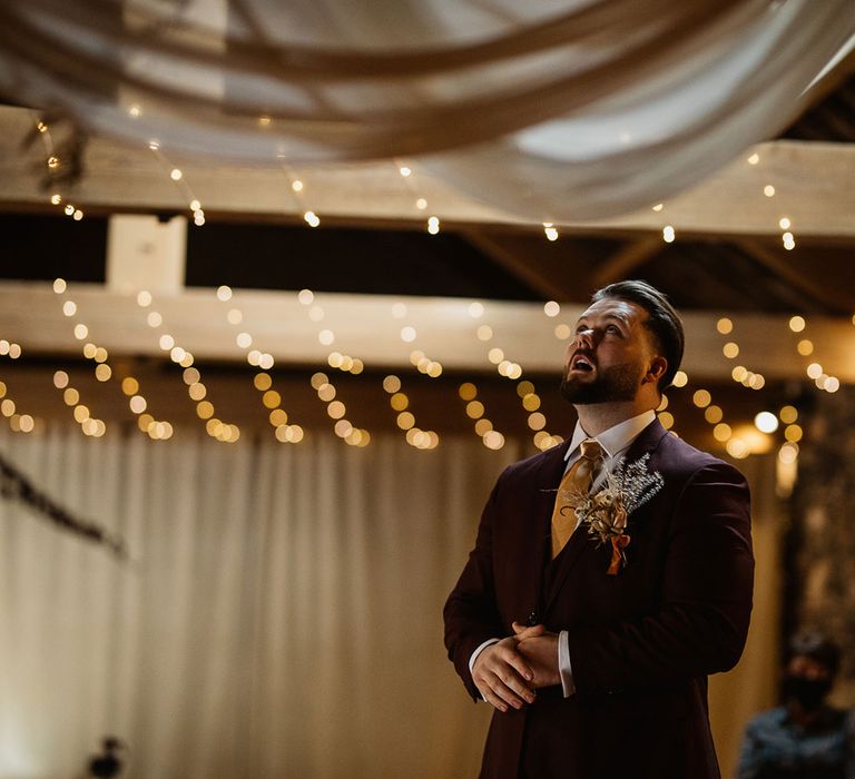 Groom in burgundy suit and gold tie waiting nervously at the altar