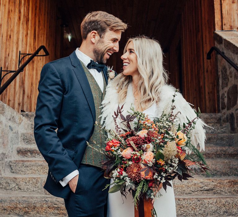 Bride wearing feathered shawl and holding bouquet looks at groom on steps