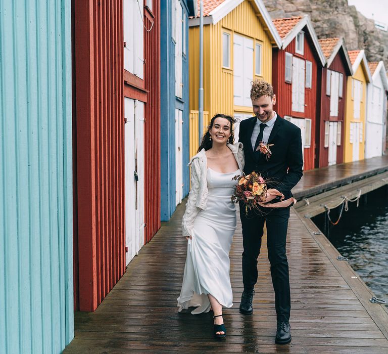 The bride and groom walking alongside colourful beach huts in Sweden