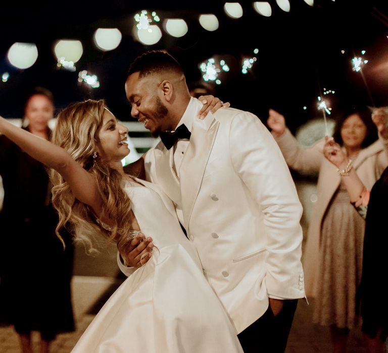 Sparkler moment with groom in a white tuxedo jacket and bride in a Justin Alexander wedding dress with long wavy hair 