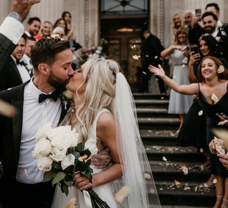 Bride & groom kiss whilst wedding guests throw confetti around them