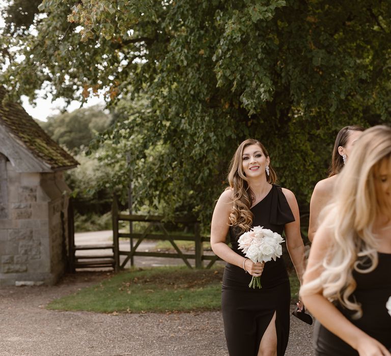 Bridesmaids in full length black dresses with long curled hair and white and blush pink rose bouquets walking outside church