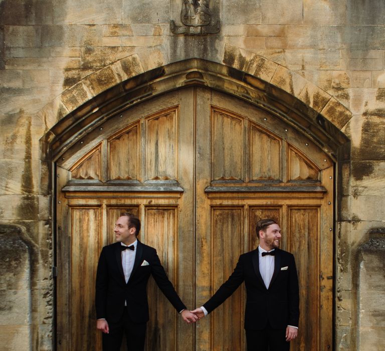 Grooms stand and hold hands outside large wooden door in Oxford