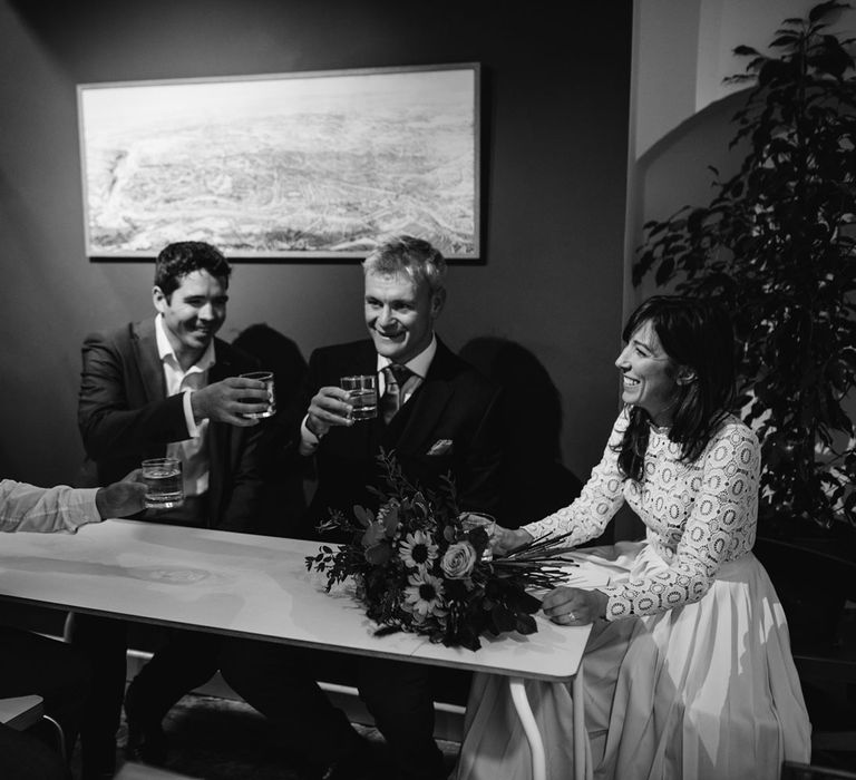 Bride in long sleeved lace top Self Portrait wedding dress raise their glasses sat in coffee shop with groom in brown woollen blazer and two friends