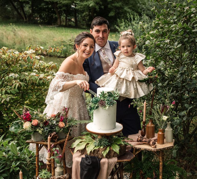 Bride & groom stand with their little girl in front of wedding cake 