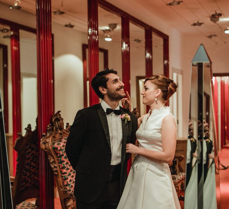 Bride and groom looking glamorous and smiling, surrounded by mirrors and red carpet in a hallway