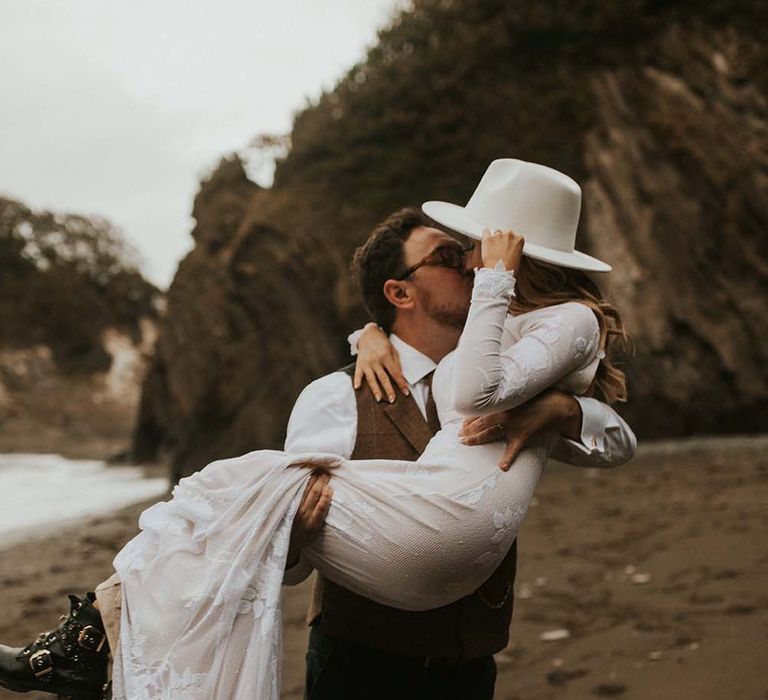 Groom picking up his bride in a boho wedding dress and fedora hat on the beach in Devon at their intimate seaside elopement 