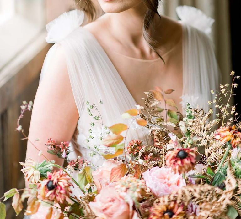 Beautiful bride with pinned bridal up do and flawless makeup sitting in a venue window holding her autumn flower bouquet 