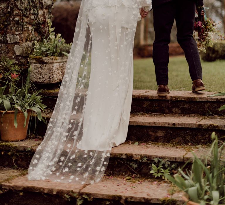 Bride in white Charlie Brear wedding dress with slit and chapel length daisy applique veil walks arm-in-arm with groom in grey tweed suit up garden steps at garden party wedding in Devon