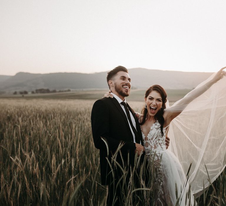 Bride and groom in a country field, the bride's veil flying in the air as she smiles