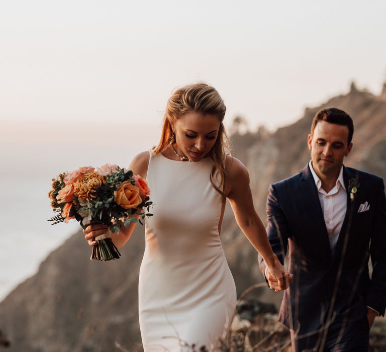 Bride holds grooms hand as they walk along the cliffside 