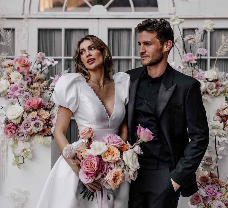 Groom in a black open shirt and tuxedo embracing his bride in a taffeta wedding dress as she holds a pink wedding bouquet 