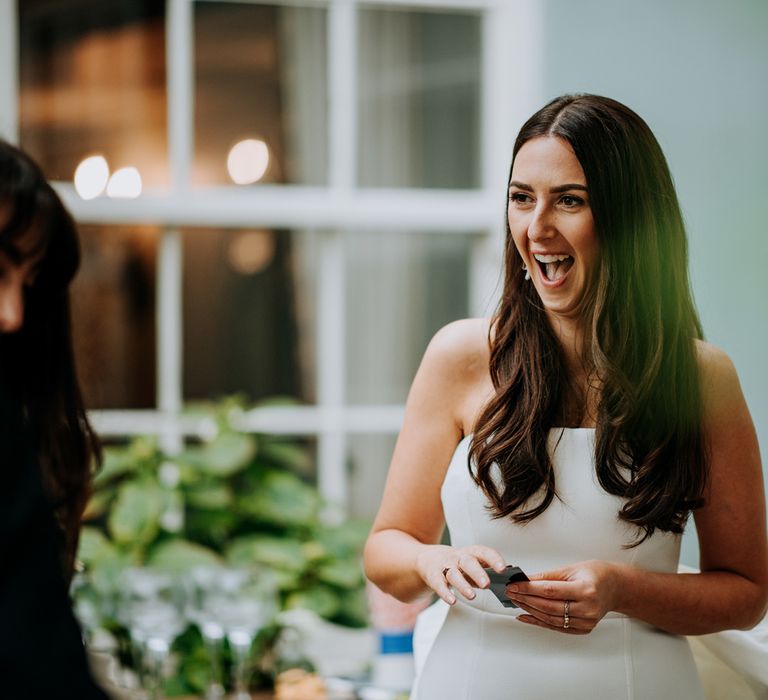 Smiling bride with long brown hair wearing white strapless Rebecca Vallance Dress stands in courtyard holding polaroids at Hotel du Vin wedding reception in Harrogate