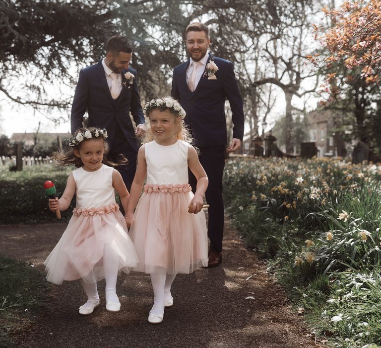 Two flower girls wearing pink tutu dresses and flower crowns walk in front of the groom and best man