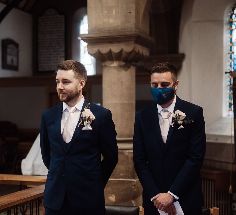 Groom and best man at the altar wearing navy blue suits with pink ties. The best man is wearing a face mask