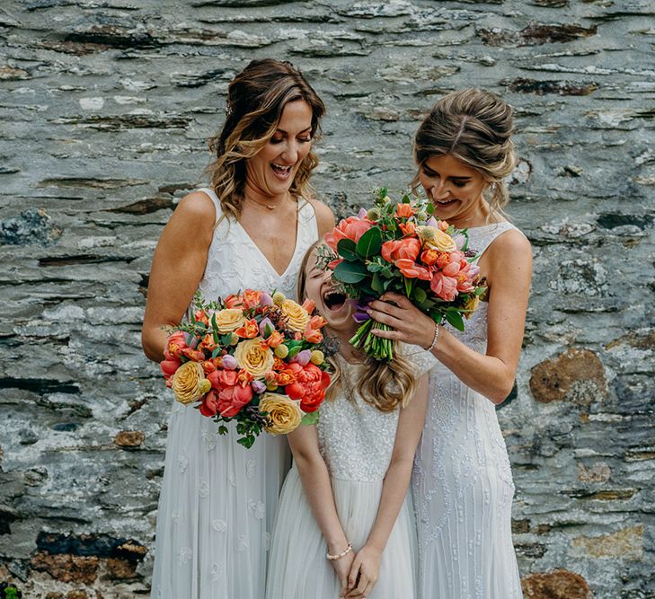 A same sex female couple laugh with their daughter during wedding portraits. They all wear white dresses and the brides' bouquets are colourful.