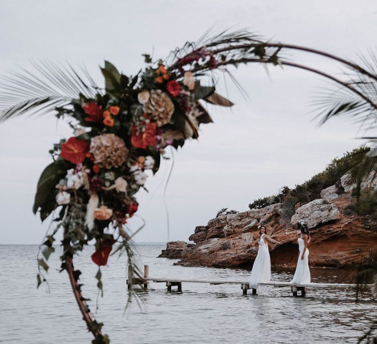 Two brides on a small wooden pier on the beach in Ibiza, with romantic red wedding floral installation