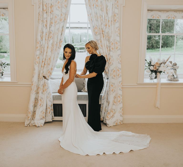 Bride with long dark curled wedding hair and white Pronovias wedding dress with train stands in bedroom at Came House Dorset with maid of honour in black one-shoulder dress before wedding