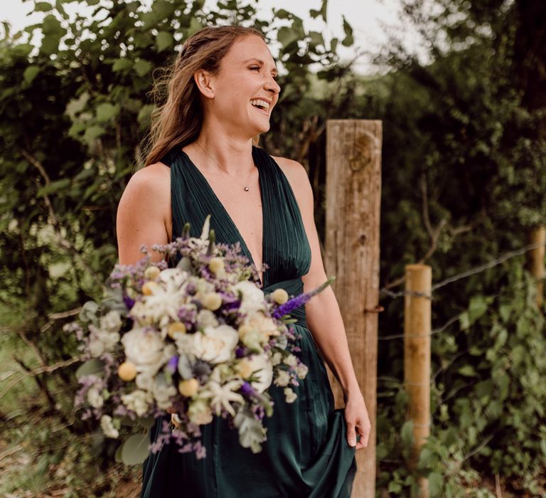 Bridesmaid in v neck forest green bridesmaid dress holds green, white and purple wedding bouquet at home farm wedding