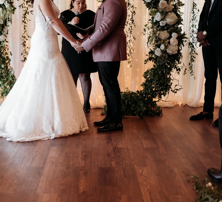 Bride & groom during wedding ceremony hold hands and face one another in front of floral archway