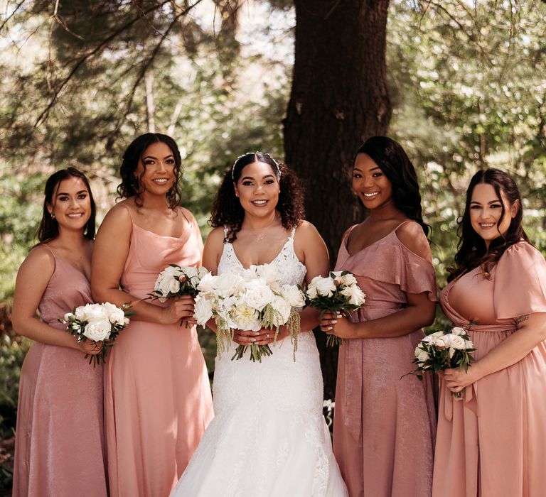 Bride stands with her bridesmaids who wears blush pink bridesmaid gowns in different styles