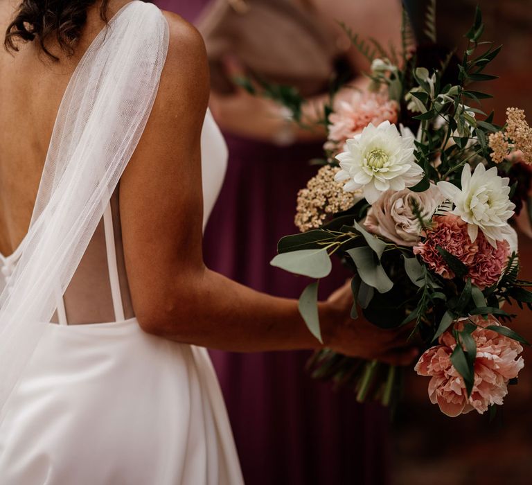 Back of bride's white cami strap Essense of Australia wedding dress with low v back, buttons and shoulder veil details as she holds white, dusky pink and green bridal bouquet for wedding at The West Mill Derby