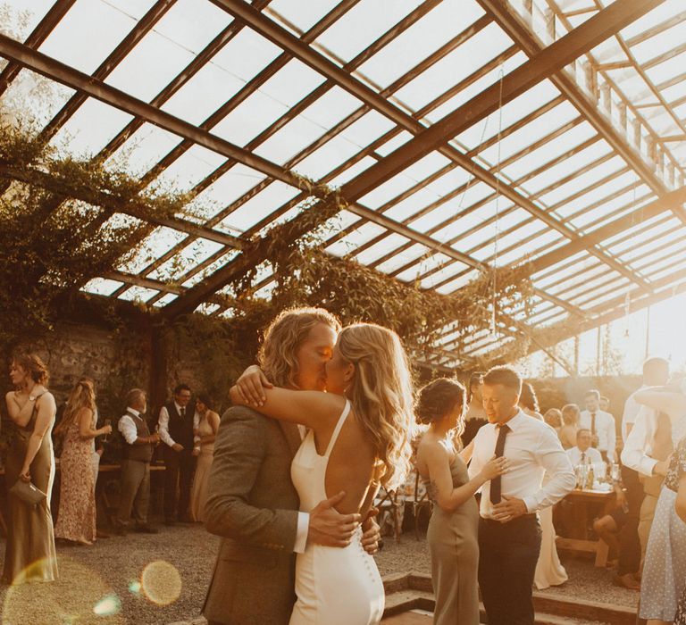 Bride in white open back Made With Love wedding dress with button details kisses groom in sage suit and brown shoes as they dance during golden hour at the glasshouse at Anran Devon