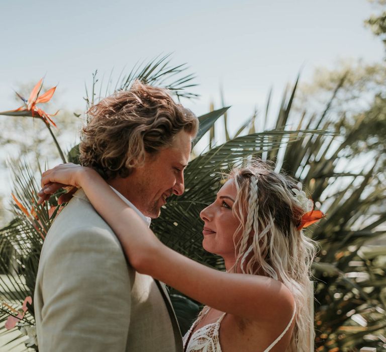 Bride in white lace boho wedding dress with blonde beach waves has arms around neck of groom in linen suit during outdoor wedding ceremony for birds of paradise wedding inspiration