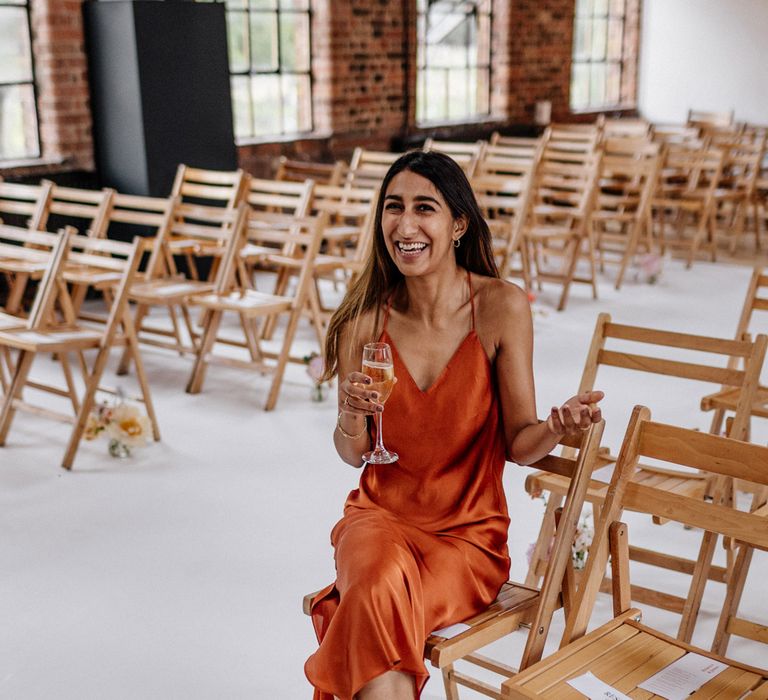 Wedding guests in burnt orange satin dress holds glass of sparkling wine whilst sat on wooden chair in wedding ceremony room at Loft Studios London