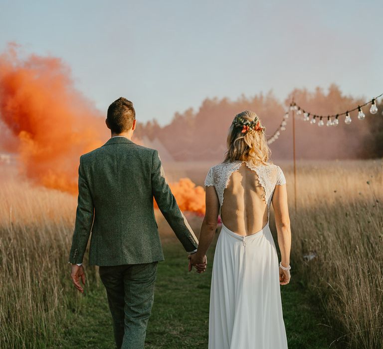 Bride in lace cap sleeved open back wedding dress with train and flowers in her hair walks towards orange smoke bomb holding hands with groom in grey herringbone suit during late summer wedding at Wellington Wood Norfolk 