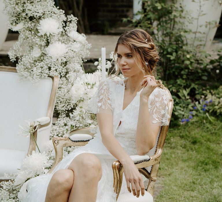 Outdoor tea party with bride in a lace wrap wedding dress sitting on an ornate white chair 