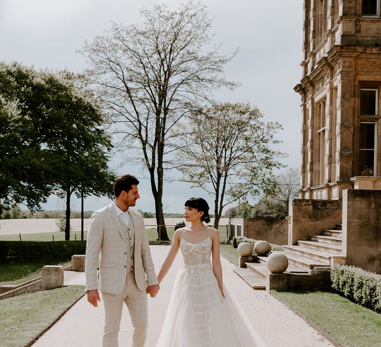 Bride in a tulle wedding dress with fitted bodice holding hands with her groom in a beige three-piece suit at Bylaugh Hall wedding venue in Norfolk 