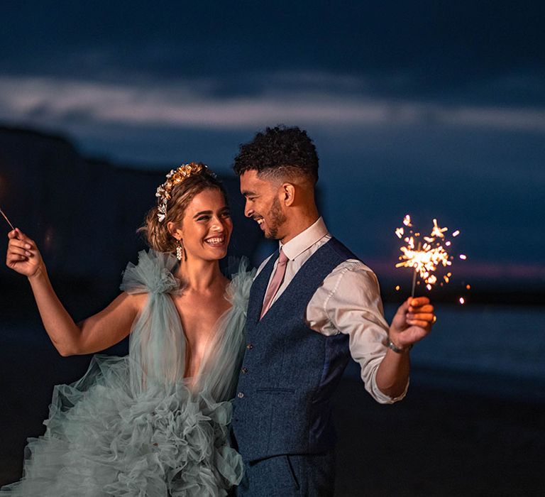 Bride in a mint green frilly wedding dress and groom a waistcoat holding sparklers on the beach 