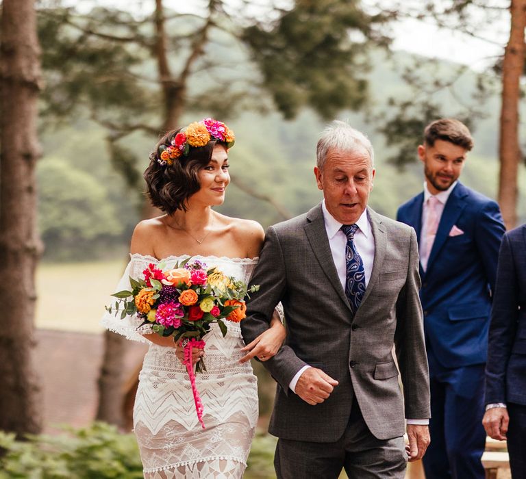 Bride in lace off the shoulder Grace Loves Lace wedding dress and flower crown carrying colourful wedding bouquet smiles whilst walking down outdoor aisle arm in arm with father of the bride in grey suit