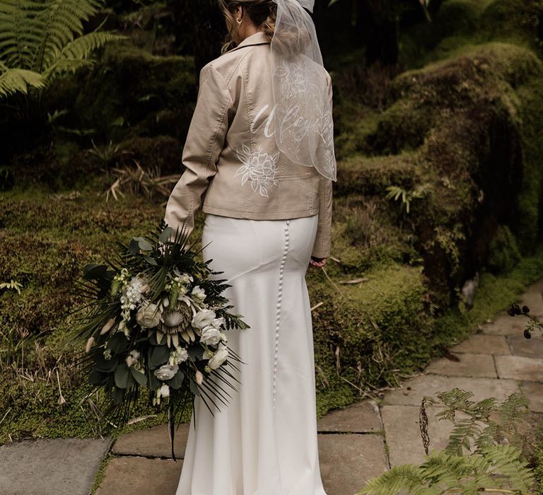 Bride in a tan leather jacket with flower motif and bow hair accessory holding a white and green bouquet 