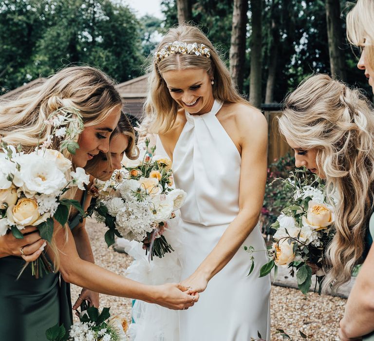 Bride in halterneck Halfpenny London wedding dress and floral headband stands with bridesmaids in green satin bridesmaid dresses as they all hold white and peach wedding bouquets