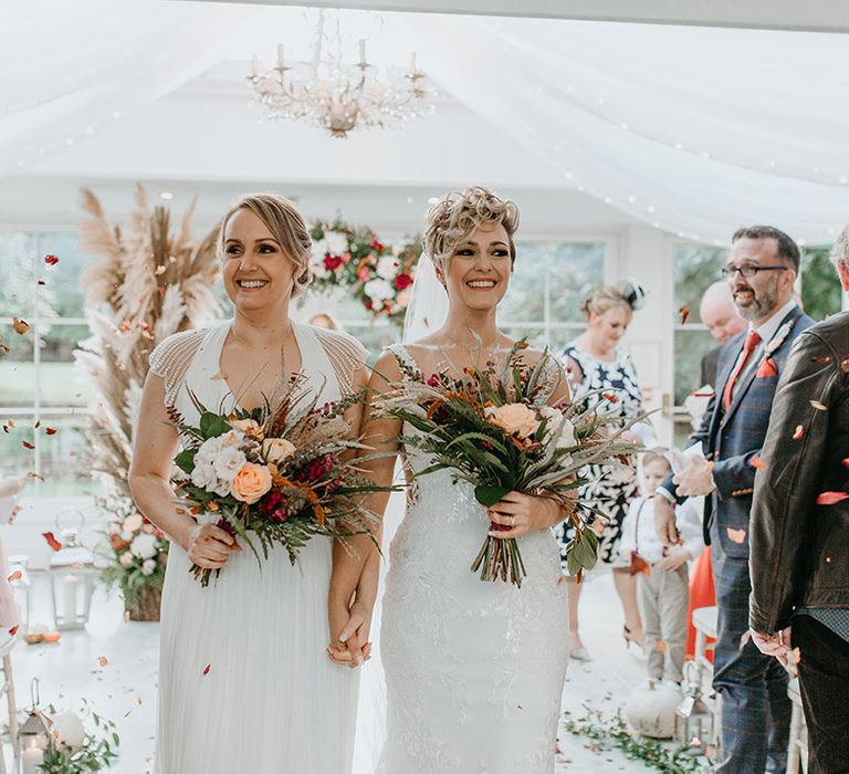 Brides walk down the aisle as they hold Autumnal bouquets 